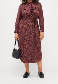 Abito in stampa paisley bordeaux con frontale arricciato, vita con cintura, maniche lunghe e chiusura con bottoni. Abbinato a una borsa nera e mocassini.