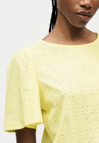 Κοντινό πλάνο ενός ατόμου που φορά μια μπλούζα από ύφασμα eyelet σε ανοιχτό κίτρινο χρώμα, με κοντά μανίκια και στρογγυλή λαιμόκοψη.