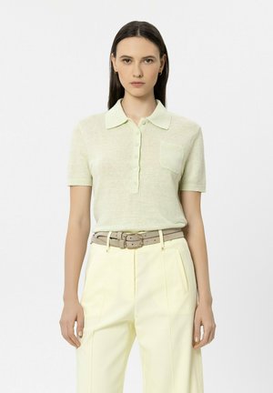 Woman wearing light green short-sleeve polo shirt tucked into high-waist pale yellow trousers with a beige double-buckle belt, standing straight.