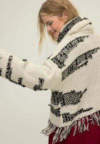 Cremfarbener und schwarzer gestrickter Cardigan mit lockerer Passform, ausgestattet mit einem hohen Kragen, Fransen am Saum und strukturierten Mustern im gesamten Design.