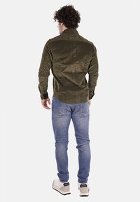 Camicia a maniche lunghe verde oliva con una sottile trama, polsini abbottonati e taglio dritto, abbinata a jeans attillati blu chiaro e sneakers.