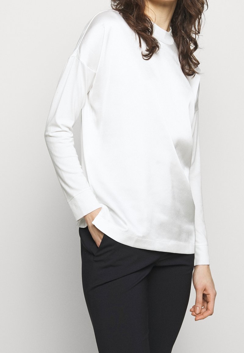 Haut blanc à manches longues avec une coupe décontractée, doté d'un col rond et de poignets, associé à un pantalon foncé, texture de tissu lisse.