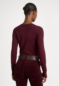 Bordový pulóverový sveter s dlhými rukávmi, rebrovanými manžetami a okrúhlym výstrihom, v kombinácii s bordovými kordurovými nohavicami a tmavo hnedým opaskom.