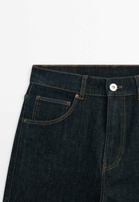 Mörkblå denimjeans har en klassisk design med knappgylf, synlig framficka och kontrasterande orange sömmar vid midjan och sömmarna.
