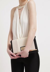 Pochette beige matelassée avec des accents texturés noirs, ferrures dorées et forme rectangulaire. Portée sous le bras avec un haut blanc drapé.