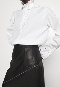 Donna che indossa una blusa bianca con pizzo sul colletto e sui polsini, abbinata a una gonna in pelle nera con zip asimmetrica.