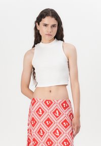 Top corto sin mangas de color blanco con un cuello alto, combinado con una falda roja con un patrón que presenta formas de diamante y motivos florales.