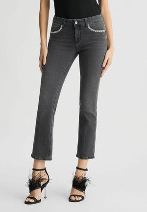 Jeans in denim grigio con una vestibilità classica, dotati di decorativi accenti in catena argentata sulle tasche. Abbinati a sandali neri con cinturini e dettagli in piuma.