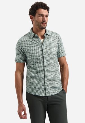 Mann mit kurzen lockigen Haaren, der ein gemustertes kurzärmliges Hemd mit Knöpfen und dunkle Hose trägt, steht mit einer Hand in der Tasche.