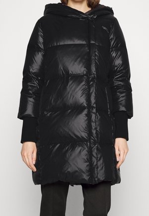 Kvinde iført en midtlængde sort dunjakke med hætte og lange ærmer over mørke bukser, stående mod en ensfarvet baggrund.