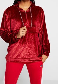 Polo en velours rouge texturé avec col zippé, capuche et cordons contrastants. Associé à un pantalon assorti rouge avec un logo.