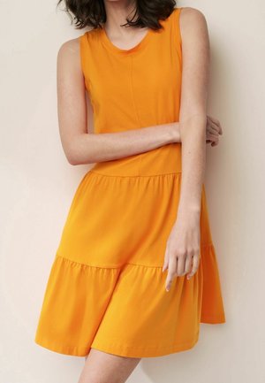 Femme portant une robe orange vif, sans manches, longueur genou, debout devant un mur clair uni, les bras croisés devant son corps.