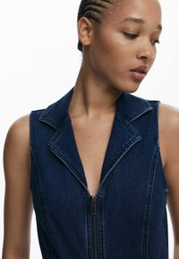 Džínový overál s hlbokým výstrihom do V, bezrukávovým dizajnom a predným zipsom. Tmavo modrá farba s viditeľnými švovými detailmi.