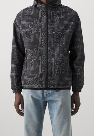 Osoba w czarnej kurtce zapinanej na zamek z wzorem złożonym z różnych napisów „Karl Lagerfeld” oraz w jasnoniebieskich dżinsach.