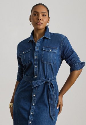 Femeie purtând o rochie bleu din denim cu nasturi, mâneci suflate, cordon în talie, cercei auriu cerc și brățară aurie pe fundal simplu.