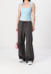 Vrouw draagt een lichtblauwe, gestructureerde, mouwloze top en losse donkergrijze broek, houdt een kleine roze handtas vast, staand op een witte vloer en achtergrond.