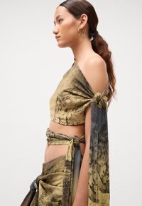 Junge Frau mit langen, zu einem Zopf gebundenen Haaren trägt ein gemustertes, asymmetrisches Cropped-Shirt und einen dazu passenden Wickelrock mit Knoten und drapiertem Stoff.