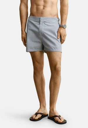 Man draagt blauw-witte gestreepte zwemshorts, een polshorloge en donkerbruine slippers, staande tegen een effen achtergrond.