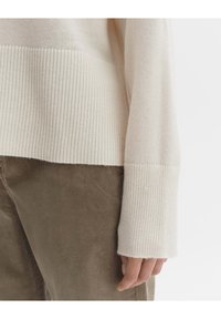 Cremefarbener, gerippter Strickpullover mit breiten Bündchen, gezeigt mit olivgrünen Cordhosen. Der Pullover hat eine strukturierte Oberfläche und einen lockeren Schnitt.