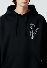 Sweat à capuche noir avec une poche avant, présentant un motif de tulipe blanche brodée avec de longues feuilles sur le côté gauche. Tissu doux, coupe décontractée.