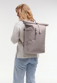 Světle šedý rolovací batoh s nastavitelnými popruhy, přední kapsou na zip a modrým, oranžovým a černým akcentem. Vyroben z odolného materiálu.