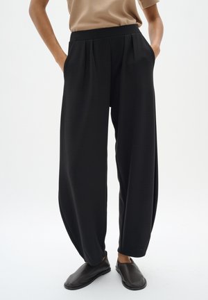 Osoba nosząca luźne czarne spodnie z plisami i kieszeniami, w zestawieniu z ciemnymi butami wsuwanymi i beżową górą, stojąca na białym tle.