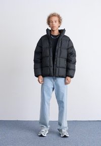 Giacca a vento nera con design trapuntato, dotata di un collo alto e tasche con zip, abbinata a jeans azzurri chiari e sneakers grigie.