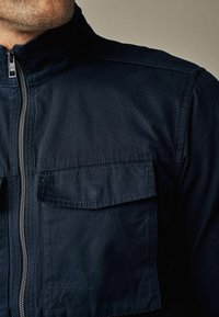 Mężczyzna ubrany w ciemnogranatową kurtkę z zamkiem z przodu i klapką na kieszeni, widoczny od szyi do połowy torsu na jednolitym tle.