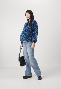 Blå zebra-mønstret bluse med puffede ærmer, parret med lyseblå klokkejeans og en sort taske. Modellen står oprejst og viser en gravid mave.