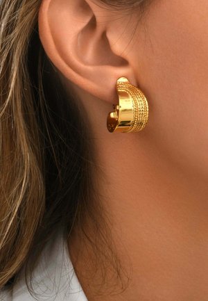 Gros plan sur une oreille portant une boucle d’oreille créole large en or texturé avec une fermeture à clip.
