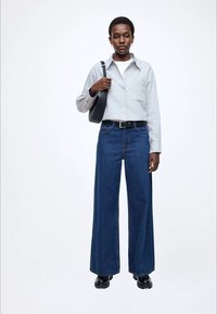 Laiade säärega tumesinised teksapüksid oranži õmblusega, kombineeritud helehallist nööbitava särgiga ja musta õlakotiga. Mustad saapad täiendavad välimust.