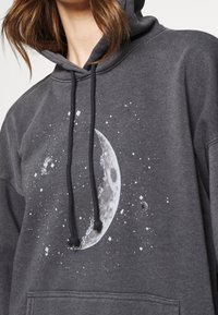 Sweat à capuche gris avec un motif frontal représentant une lune avec des éclaboussures. Il a une coupe ample, des cordons de serrage et une texture douce.