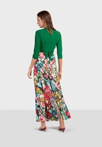 Top verde avvolgente con maniche a tre quarti abbinato a una gonna maxi fluida con motivo floreale colorato. Dettaglio di cravatta delicato sulla parte posteriore.