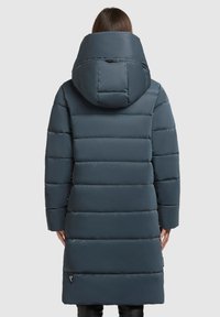 Duga pufasta jakna u tamnoplavoj boji, s visokim ovratnikom i prevelikom kapuljačom. Ima kvadratne panele i elegantnu, glatku teksturu.