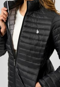 Černá péřová bunda se zipovým uzávěrem, vodorovným prošíváním, vysokým límcem a malým bílým logem na vnitřní podšívce hrudníku v šedé barvě.