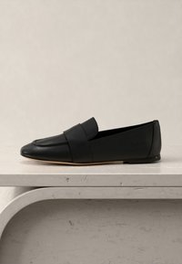 Μαύρο δερμάτινο μοκασίνι χωρίς κορδόνια με χαμηλό τακούνι, στρογγυλεμένη μύτη και φαρδιά λουρί στο πάνω μέρος, τοποθετημένο σε ανοιχτό γκρι επιφάνεια.