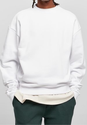 Person trägt ein schlichtes weißes, übergroßes Sweatshirt über einem cremefarbenen Langarmshirt mit dunkelgrünen Hosen, Hände in den Taschen, neutraler Hintergrund.