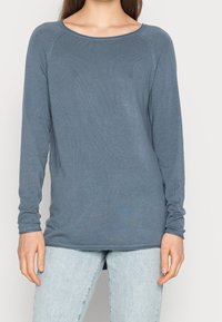 Langärmliges blaues Oberteil aus geripptem Stoff, mit einem runden Halsausschnitt und einem etwas längeren Rückensaum. Kombiniert mit hellen Jeans.