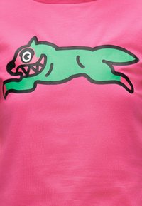 Πράσινος καρτούν σκύλος με μυτερά δόντια και μεγάλα μάτια, που πηδά προς τα δεξιά, πάνω σε φωτεινό ροζ ύφασμα.