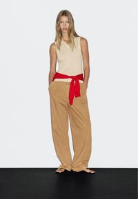 Beige ærmeløs top parret med løse, lysebrune, højtaljede bukser. En rød sweater er bundet om livet. Sorte flip-flops.