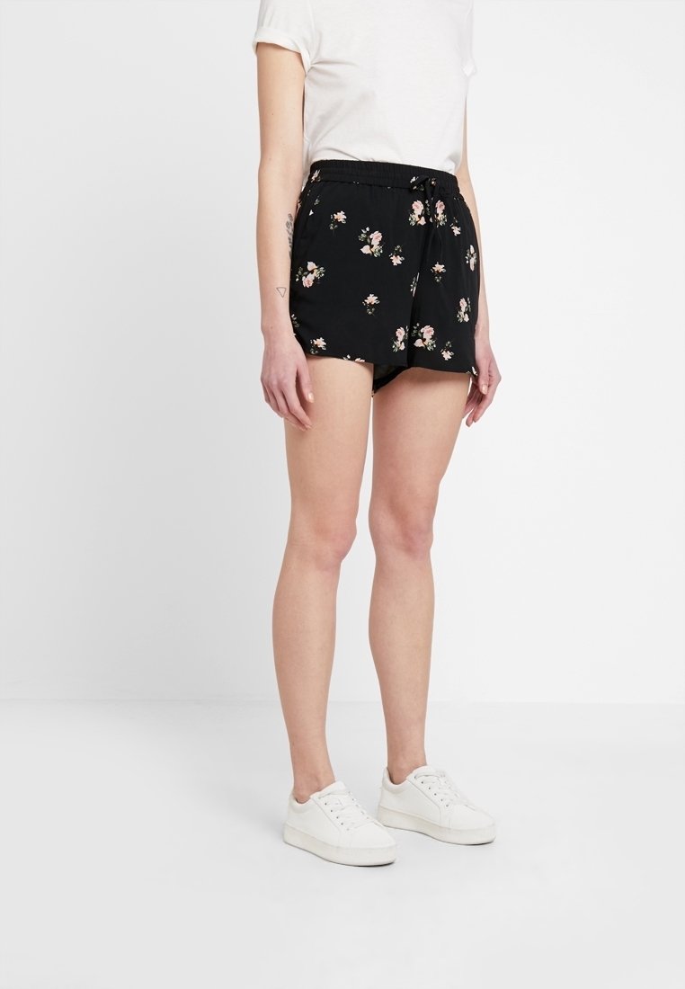 Personne portant un short noir à fleurs, un t-shirt blanc et des baskets blanches, debout devant un fond blanc uni.