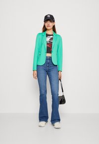 Mätovo zelený blazer, čierny priesvitný crop top, modré zvonové džínsy, biele tenisky a čierna čiapka. Drží malú čiernu kabelku.