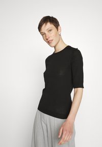 Black short-sleeve knit top with a round neckline and subtle shoulder details, paired with light gray pleated trousers. Smooth texture.