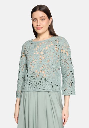 Kvinde iført en lyseblå-grøn hækleblomstret langærmet top over et matchende ensfarvet nederdel, stående mod en ensfarvet baggrund.