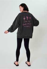 Übergroßer Sweatshirt in Anthrazit mit pinkem grafischen Text und Illustrationen auf der Rückseite. Kombiniert mit schwarzen Leggings und Flip-Flops.