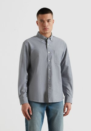 Młody mężczyzna w jasnoszarej koszuli na guziki z długimi rękawami i niebieskich dżinsach, stojący na tle białej ściany.