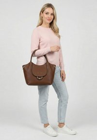 Femme portant un pull rose et un jean clair, tenant un grand sac à main en cuir marron avec rabat et détail anneau en métal.