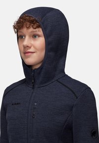 Giacca in pile con cappuccio blu navy, dotata di una mezza cerniera e dettagli neri. Presenta un tessuto testurizzato e il logo Mammut sul petto e sulla manica.