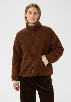 Donna con giacca in pile marrone con zip, mani nelle tasche, maniche beige del maglione visibili e pantaloni di velluto a coste marrone scuro.