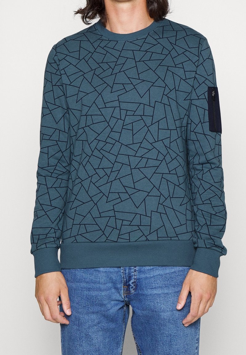 Sweatshirt turquoise avec un motif géométrique noir, des poignets et un ourlet côtelés. Comprend une poche zippée sur la manche gauche. Texture de tissu douce.